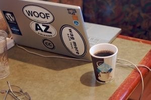 A sticker-covered laptop computer and a mug of coffee sit on a coffeehouse table.