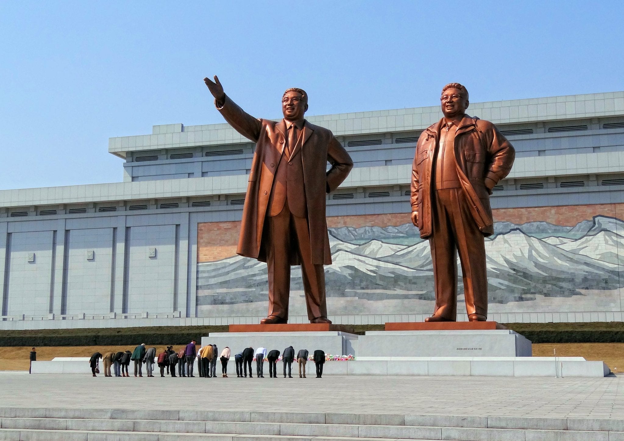 Visitors bowing in a show of respect for North Korean leaders Kim Il-sung and Kim Jong-il on Mansudae (Mansu Hill) in Pyongyang, North Korea; image by Bjørn Christian Tørrissen (Own work), CC BY-SA 3.0, via Wikimedia Commons, no changes.
