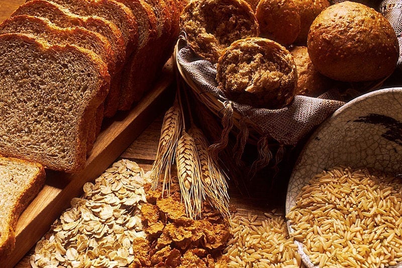 A sliced loaf of brown bread, a basket of rolls and 4 types of grains sit on a wooden table.