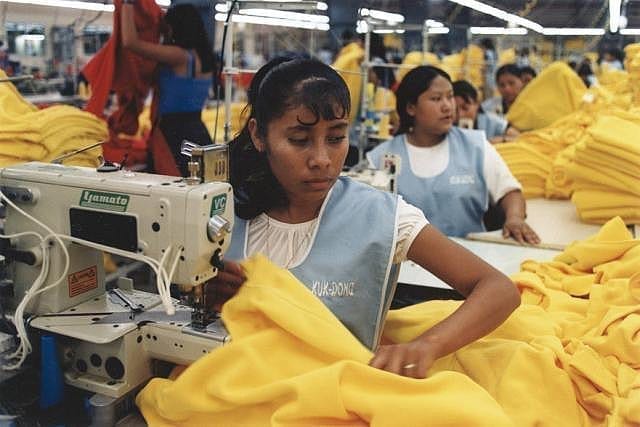 A woman with dark skin sews clothing in a sweatshop factory, surrounded by identically dressed women also sewing clothes.