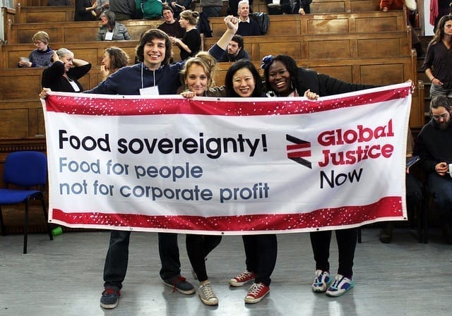 A diverse array of attendees at a 2015 UK food sovereignty gathering hold a banner. It says Food sovereignty! Food for People, not for corporate profit / Global Justice Now.
