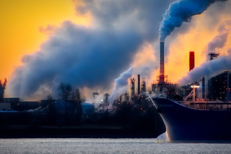 Thick blue smoke rising from an industrial complex in front of a glowing sky.