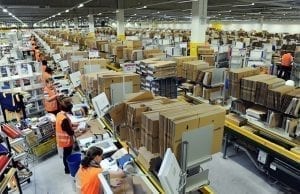 Amazon workers at a conveyor belt in a cavernous warehouse, shelves of goods as far as the eye can see.