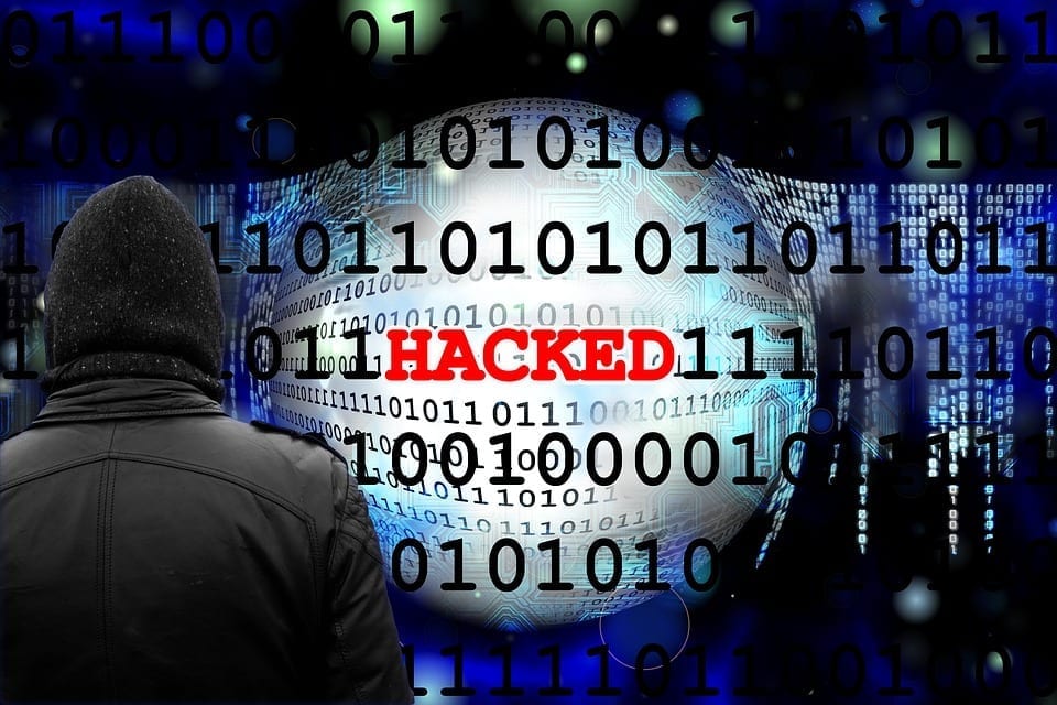 A black-clad figure faces away from the camera. In the background, the word HACKED is spelled out in red letters against a backdrop of black ones and zeroes.