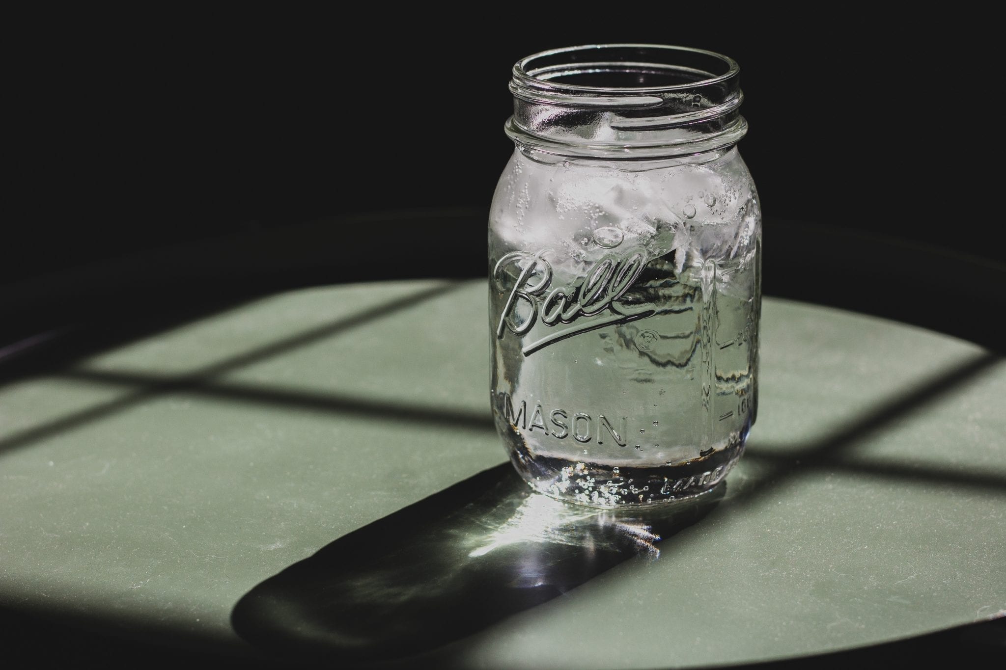 Sunlight filters through a Ball jar of clear, icy water sitting on a grey surface with a dark background.