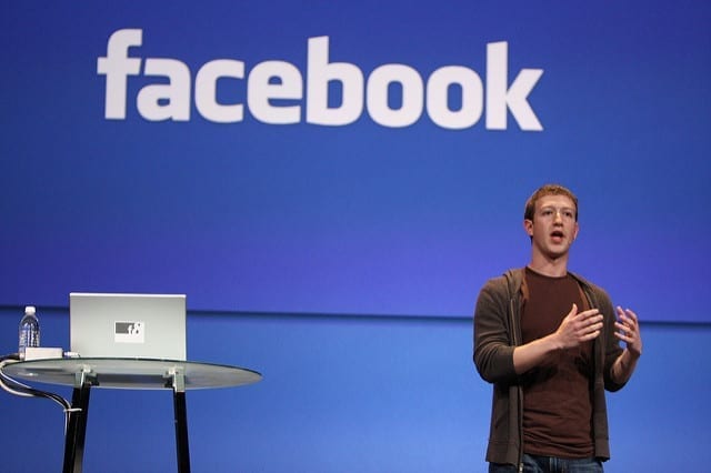 Mark Zuckerberg stands to speak in front of a blue background with the Facebook logo, alongside a small take with water and a laptop computer.