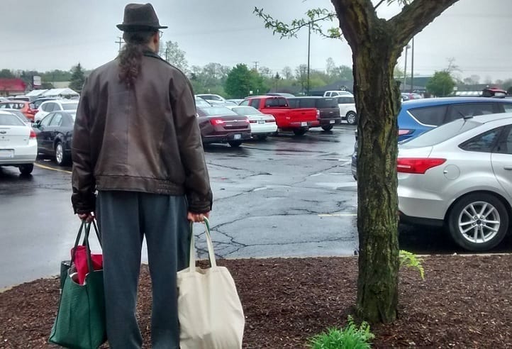A long-haired man in a leather jacket, toting cloth bags full of groceries, faces away from the observer as he scans a parking lot.
