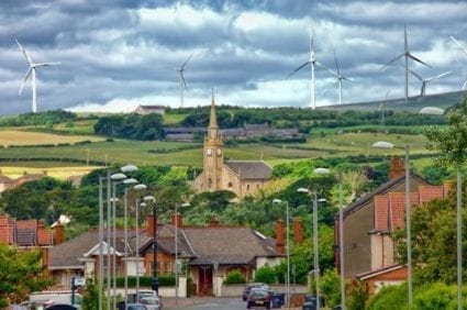Wind turbines against a cloudy sky serve as a backdrop for a street and buildings in a small, rural town.