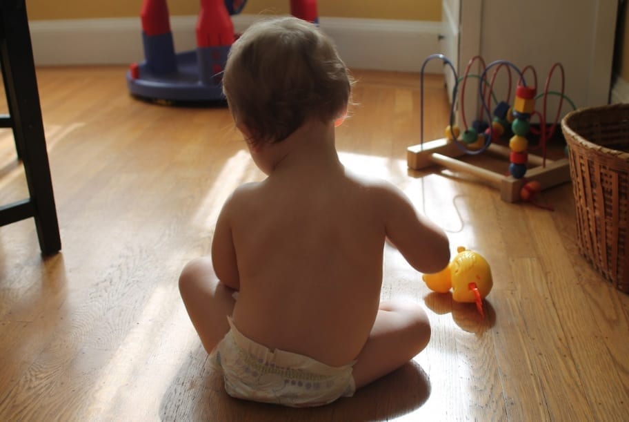 A caucasian baby, seen from behind, playing on a wooden floor with a few toys in the background.