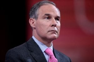 A portrait of an unsmiling Scott Pruitt, wearing a grey suit with a red tie, against a somewhat blurry red and black background.