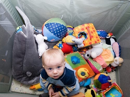 A caucasian baby in a playpen, seen from above, surrounded with stuffed animals and other soft toys.