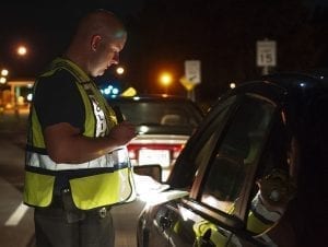 A police officer with a flashlight peers into the driver's side window of a car at night.