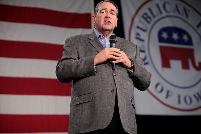 Mike Huckabee, wearing a suit, standing in front of a large American flag and a Republican elephant seal, addressing an unseen audience.