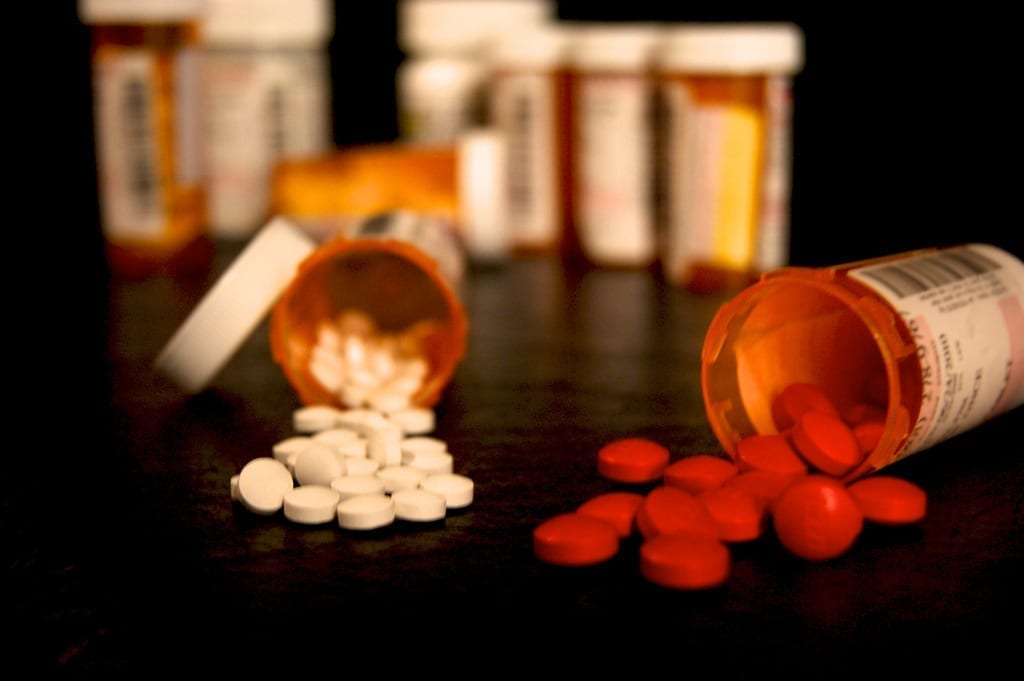 Two prescription pill bottles lie on their sides, spilling out reddish and white pills. Other pill bottles sit in the distance.
