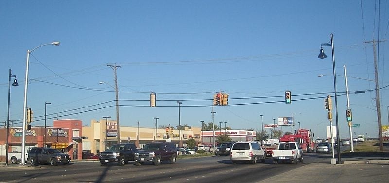 A suburban street view with wide traffic lanes, an intersection with a traffic light, and a strip mall.
