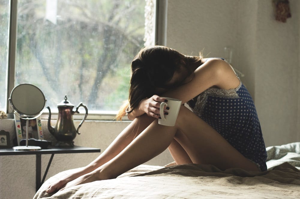 Sad woman in bed holding coffee mug with her head on her knees; image by Asdrubal luna, via Unsplash.