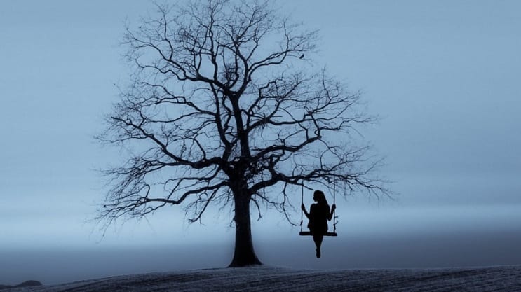 A lone figure sits on a swing that hangs from a bare tree, all in dark silhouette against a grey background.