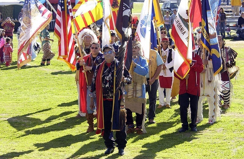 Native Americans in colorful regalia stand in loose formation, posting their colors: various flags of the United States, military branches, states and Native nations.