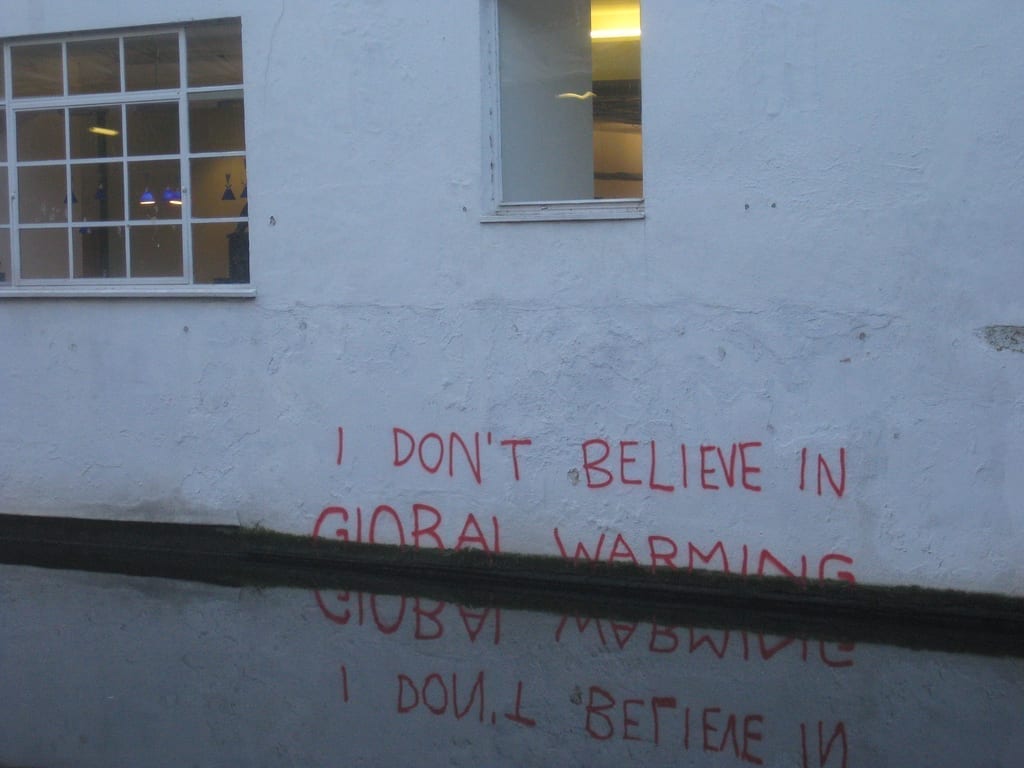 On a white wall, partially obscured by rising flood water, are the scrawled words "I Don't Believe in Global Warming."
