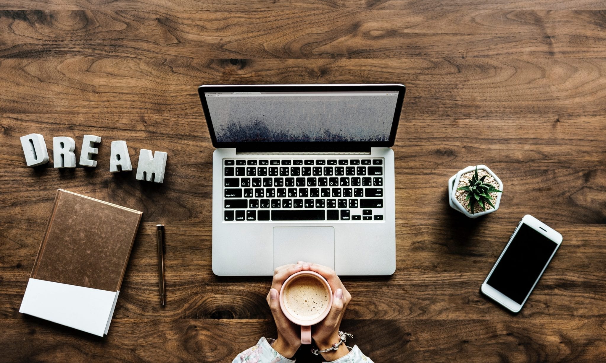 Woman sitting at table with laptop, coffee, and a sign saying “Dream;” image by Rawpixel, via Unsplash.com.