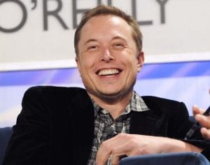 Elon Musk, grinning and at ease, wearing a dark suit and a plaid shirt with an unbuttoned collar.