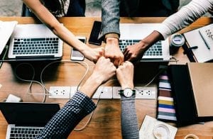 Team of five bumping fists over laptop computers; image by Rawpixel, via Unsplash.com.