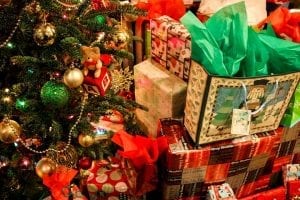 Christmas gifts in bags and boxes piled high near a festively decorated Christmas tree.