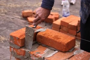 A man's hand spreads mortar with a trowel as he builds a wall from common red bricks.