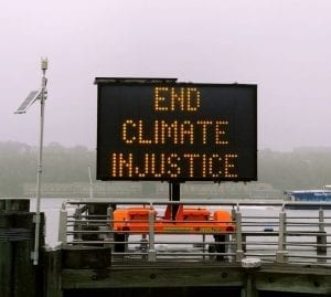 Lighted sign saying “End Climate Injustice.” Image by Jon Tyson, via Unsplash.com, cropped to reduce visible sky.