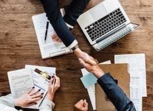 People shaking hands over a table with paperwork and a laptop computer; image by Rawpixel, via Unsplash.com.