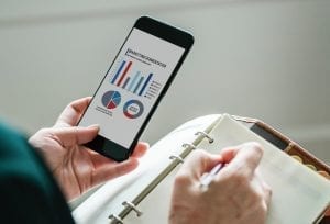 Man looking at marketing graphs on a cellphone while making notes; image by Rawpixel, via Unsplash.com.