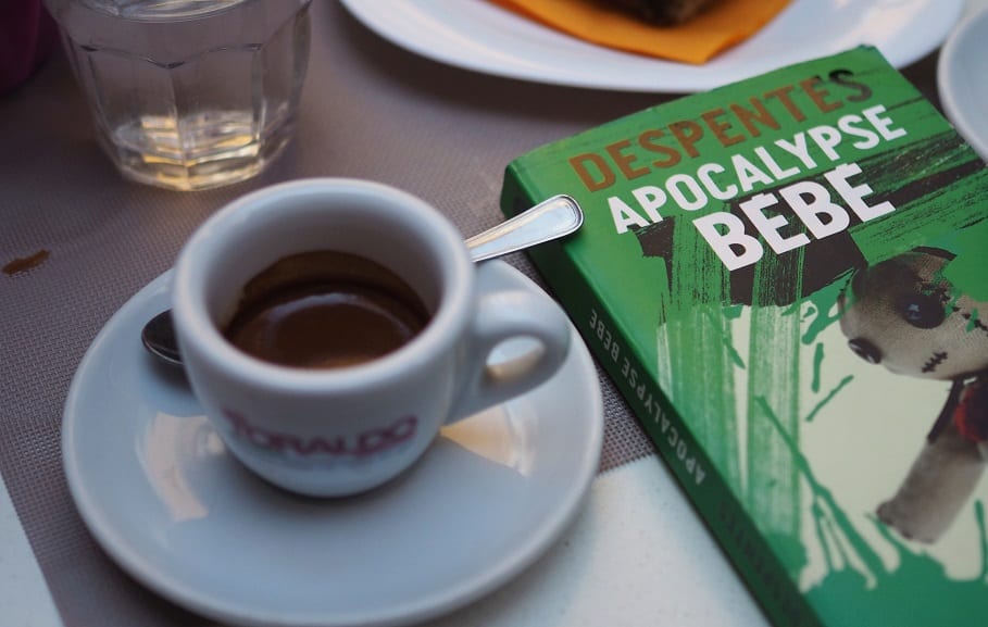 A small, white mug of black coffee on a table next to the book Apocalypse Bébé.
