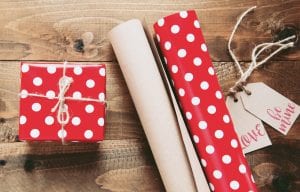 Box wrapped in red paper with white dots sitting next to wrapping paper and gift tags saying “Love” and “Be mine.” Image by Miroslava, via Unsplash.com.