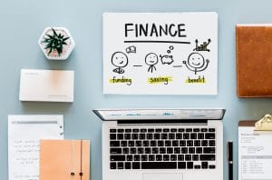 Table with laptop, plant, pen, notepads, and a sign reading “Finance: Funding, Saving, Benefit.” Image by Rawpixel, via Unsplash.com.