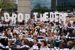 A crowd of people, bound by paper chain links, hold up letters that spell out "Drop The Debt."