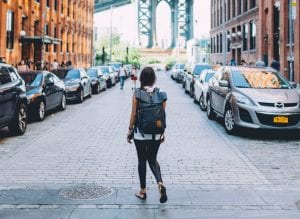 Woman wearing backpack standing on a cobblestone road; image by Brevitē, via Unsplash.com.