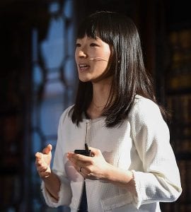 Marie Kondo speaking in front of a dark background, wearing a white cardigan and a microphone.