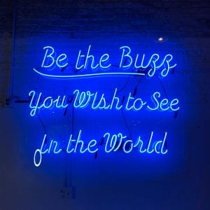 Neon blue “Be the buzz you wish to see in the world” sign; image by Tanya Santos, via Unsplash.com.