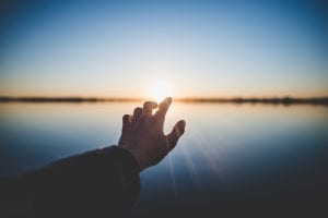 Hand reaching toward sunrise over a lake; image by Marc-Olivier Jodoin, via unsplash.com.