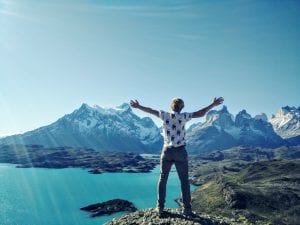 Man with hands in the air, joyfully looking at water and mountains; image by Philippe Siguret, via Unsplash.com