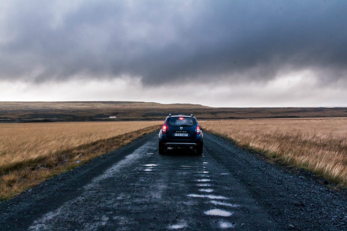 Car driving down dirt road through fields of brown grass, storm clouds in the sky; image by Kévin Langlais, via unsplash.com.