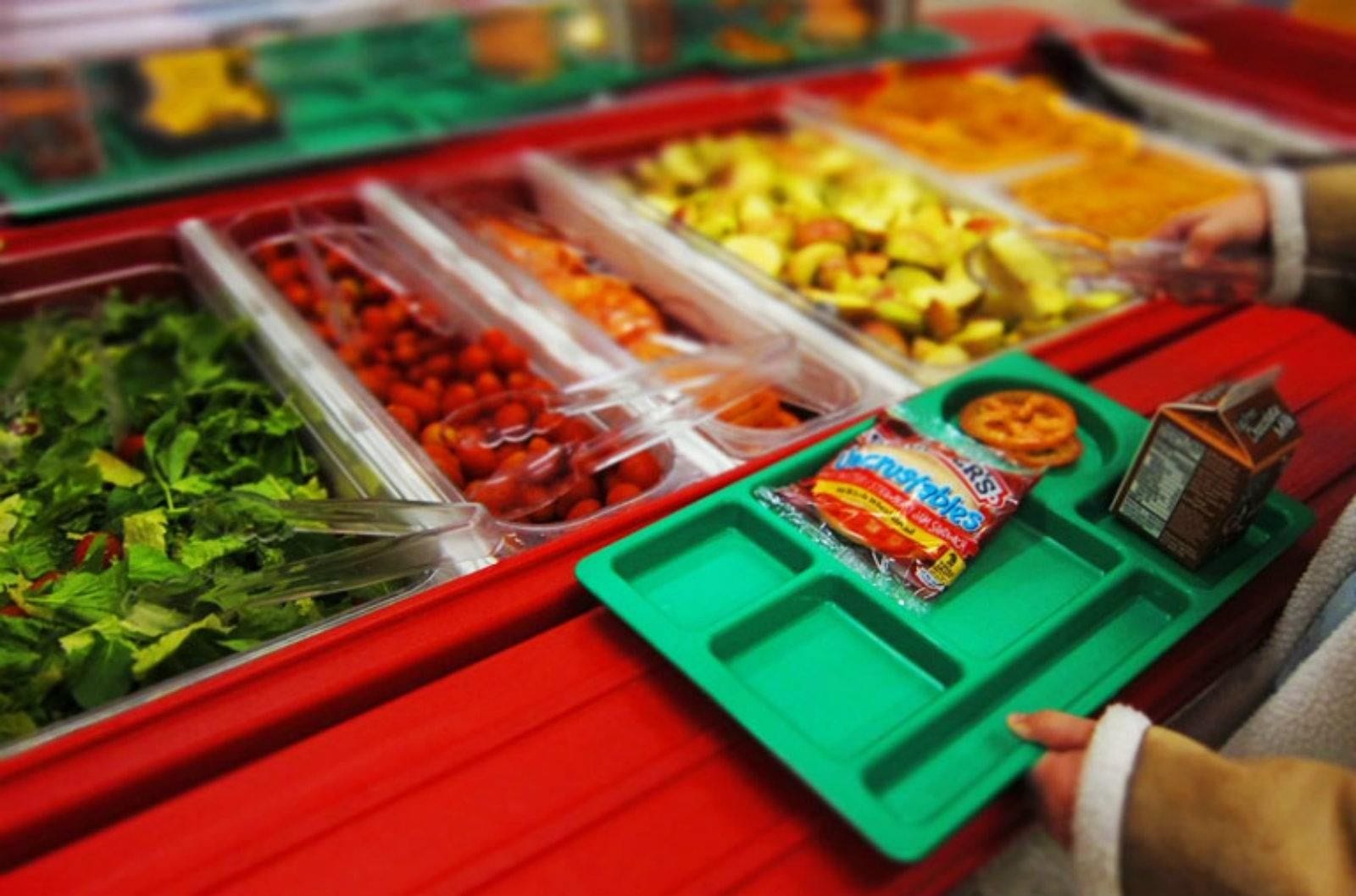 A young person carrying a tray chooses among many brightly colored fruit and vegetable options in a cafeteria setting.