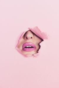 Woman’s nose and mouth, visible through a hole torn in pink paper background; image by Ian Dooley, via unsplash.com.