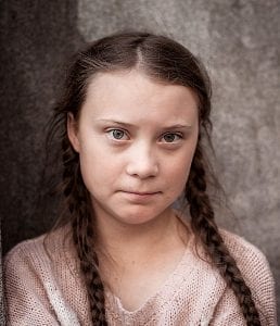 A serious-looking teenage girl with braided hair, wearing a pale colored sweater against a dappled grey background wall.