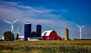 A classic red barn, surrounded by agricultural fields, silos, and wind turbines.