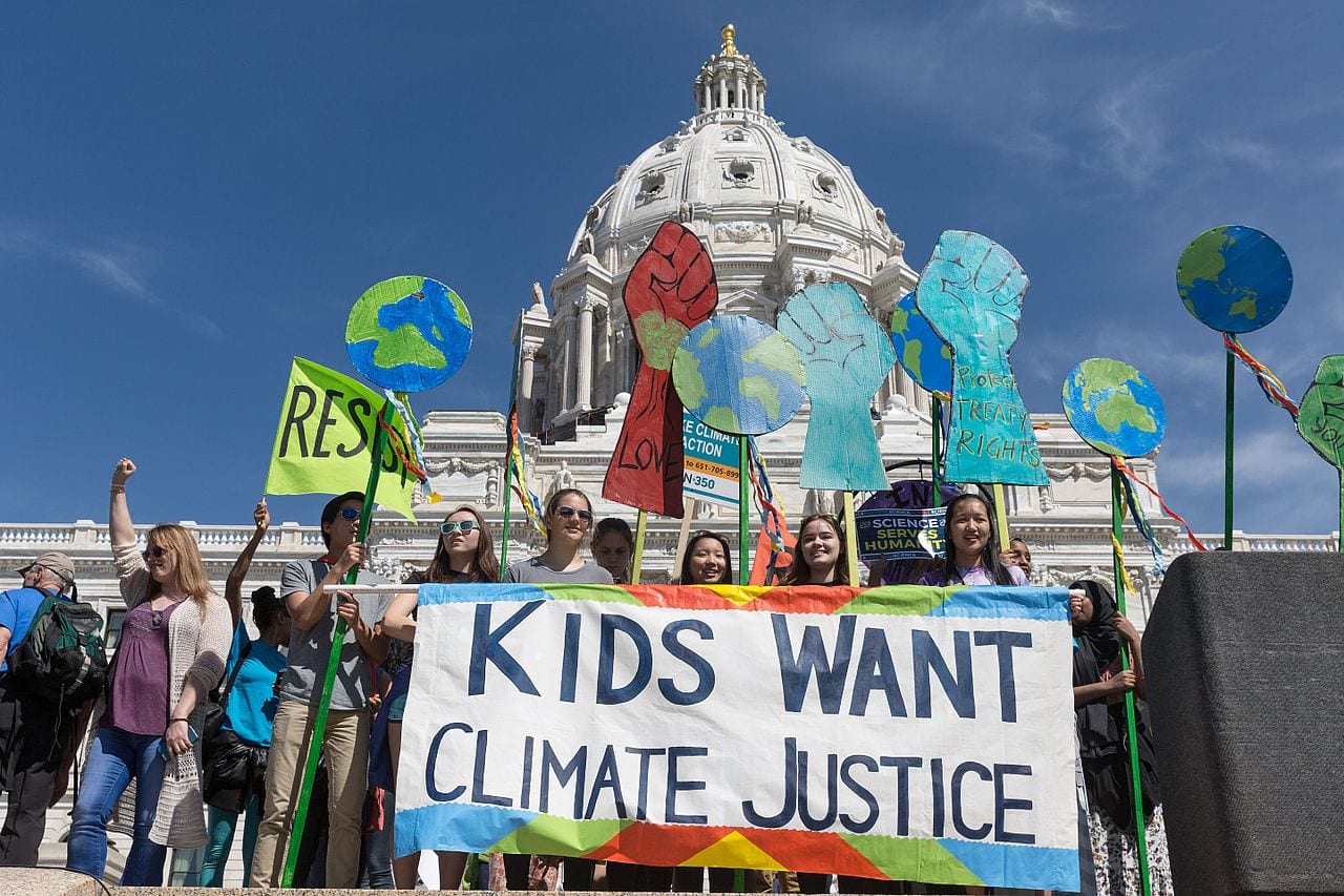 Young people holding protest signs and a banner reading "Kids Want Climate Justice" in front of of a domed building on a clear day.
