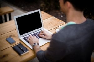 Man seated at an outdoor table working on a laptop; image by Alejandro Escamilla, via unsplash.com.