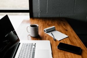 Laptop, smartphone, pad of paper and pen, and cup of coffee on a woodgrain table; image by Andrew Neil, via unsplash.com.