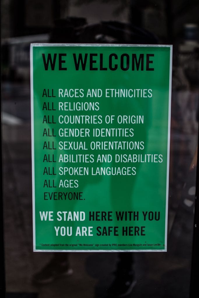 Green sign with white lettering that reads “We welcome all races, ethnicities, religions, countries of origin, gender identities, sexual orientations, abilities and disabilities, spoken languages, ages, everyone. We stand here with you. You are safe here.” Image by Brittani Burns, via unsplash.com.