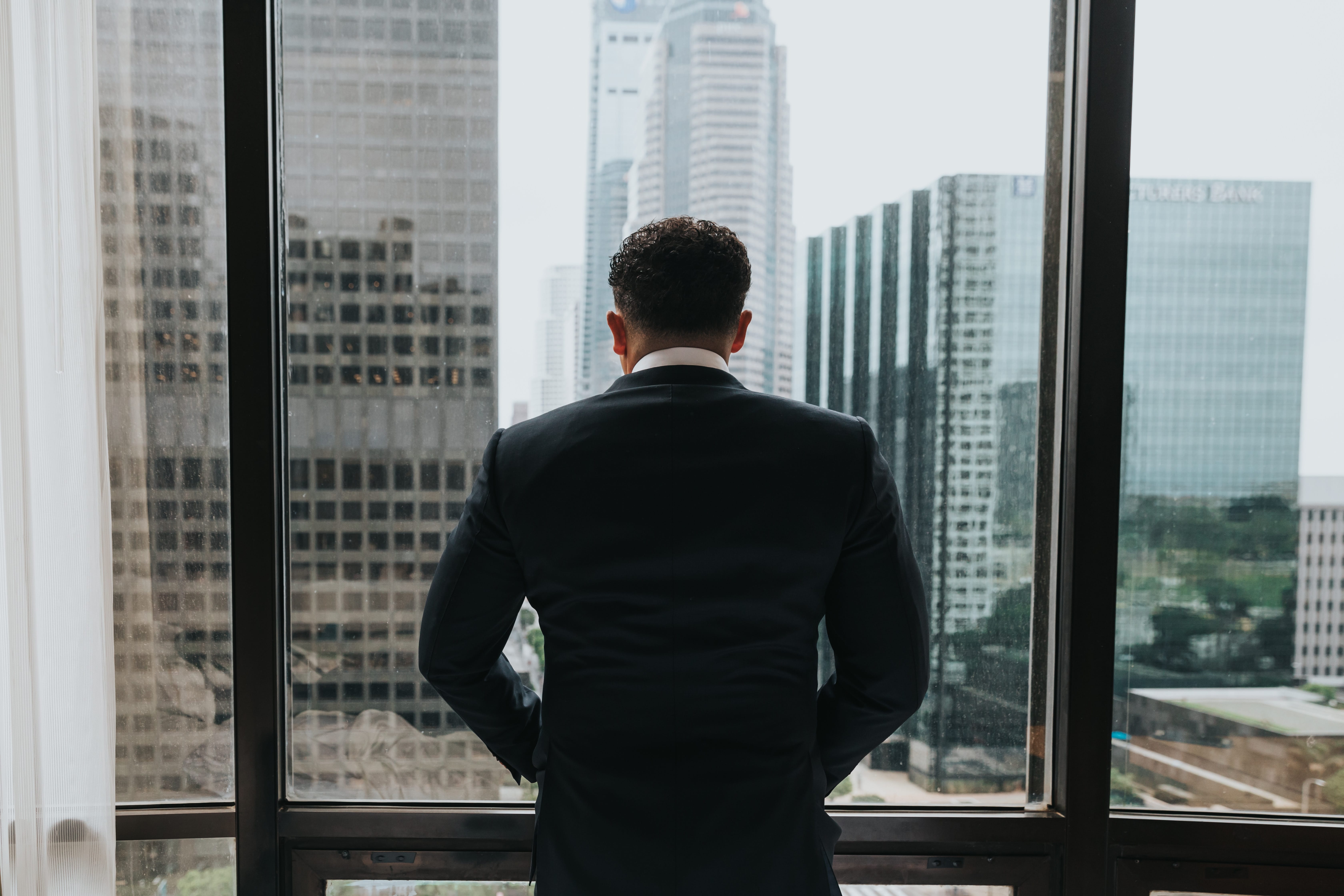 Man in suit, hands in pockets, looking out window of office building; image by Nathan Dumlao, via Unsplash.com.